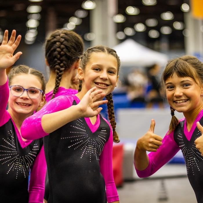Four young gymnasts at a sports arena, three visible and smiling at the camera. They are wearing matching black and pink leotards with sparkling designs. One girl with glasses is waving, another is smiling broadly with braided hair, and the third is giving a thumbs-up.