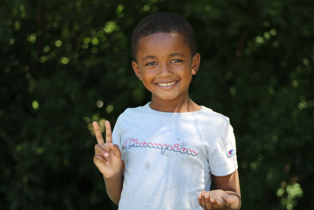 A young boy with a bright smile is standing outdoors against a backdrop of lush green foliage. He is wearing a light gray T-shirt with the word 'Champion' written across the front. The boy is holding up his left hand making a peace sign with his fingers, and his right hand is slightly open. His cheerful expression and the vibrant greenery create a joyful and welcoming scene.