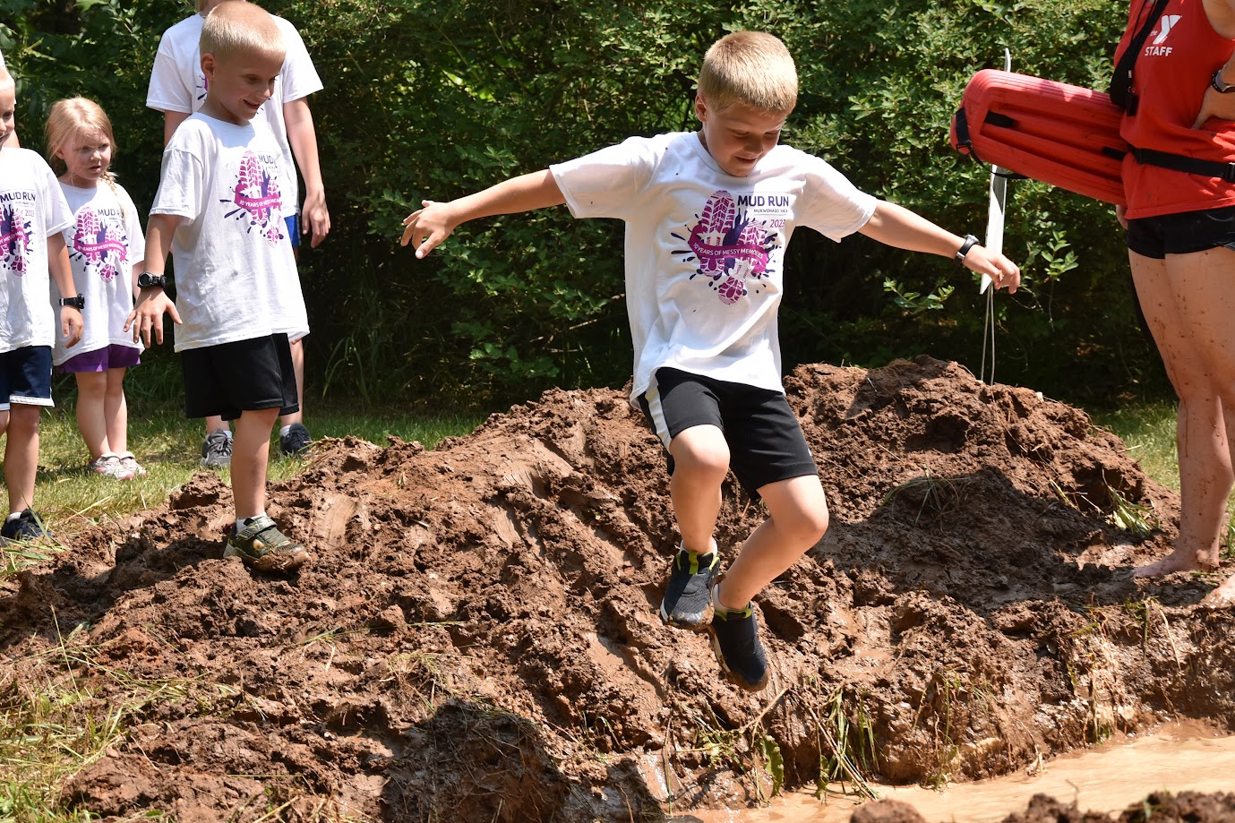 A group of children wearing white t-shirts with a purple 'Mud Run' logo, standing on a muddy track. One boy is seen jumping energetically into the mud while others watch in anticipation. A staff member wearing a red shirt stands nearby, holding a safety float.