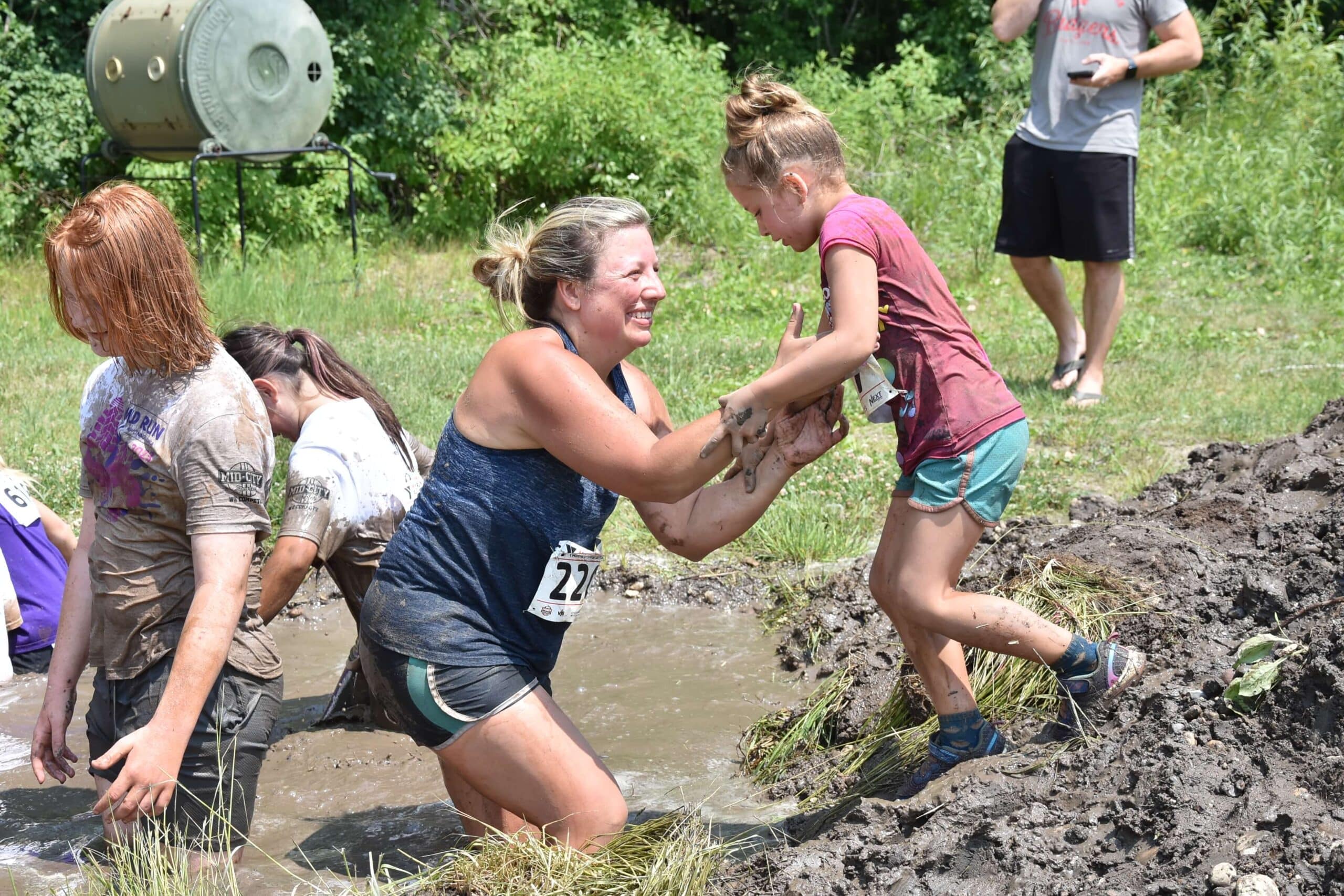 A woman in a blue tank top helps a young girl in a maroon shirt and blue shorts climb out of a muddy pit during a mud run. Both are smiling and covered in mud. Other participants are seen in the background, also navigating the muddy course. The atmosphere is lively and playful.