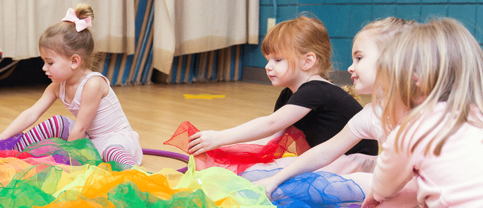Three young girls participating in a dance class indoors. They are seated on the floor, interacting with a colorful, rainbow parachute. The girls are dressed in casual dance attire, with the central child in a white leotard and striped leggings, focused on stretching the parachute
