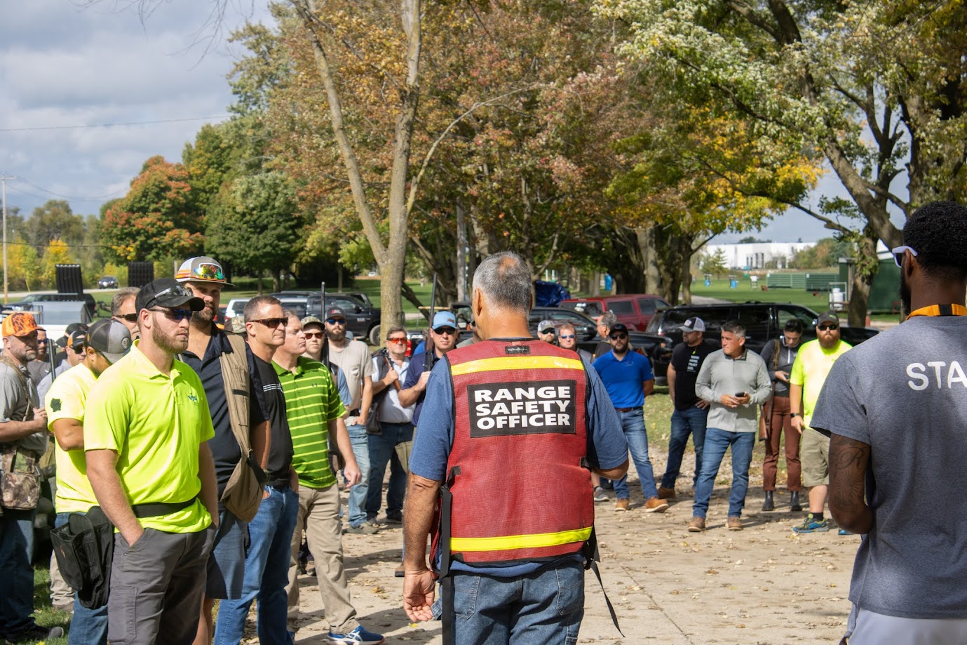 A diverse group of men, attentively listening to a range safety officer who is wearing a red vest labeled 'RANGE SAFETY OFFICER'. The setting is an outdoor park with scattered trees and greenery, under a partially cloudy sky. Some attendees are in casual attire, while others wear uniforms or safety gear