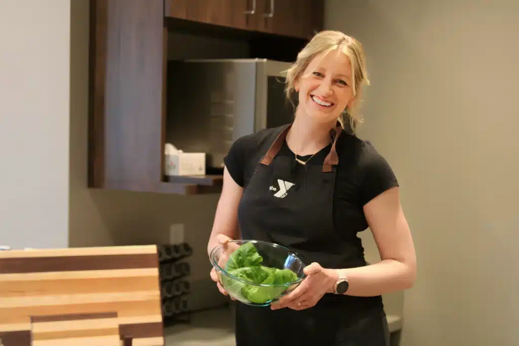 A woman with a joyful expression stands in a modern kitchen, holding a clear bowl filled with fresh green leaves. She is wearing a black apron with the YMCA logo on it. The kitchen setting is sleek and minimalistic, featuring wooden furnishings and neutral tones.
