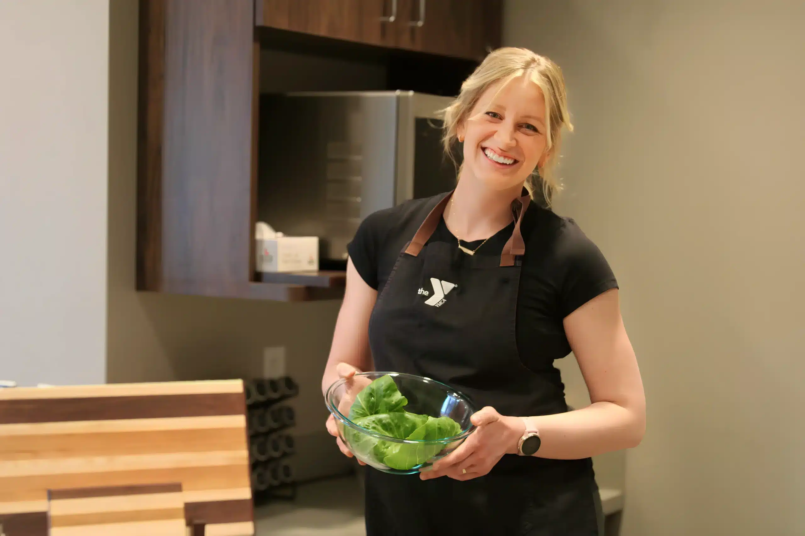 A woman with a joyful expression stands in a modern kitchen, holding a clear bowl filled with fresh green leaves. She is wearing a black apron with the YMCA logo on it. The kitchen setting is sleek and minimalistic, featuring wooden furnishings and neutral tones.