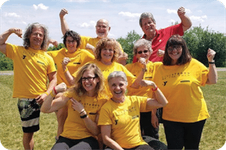 A group of people standing outside on grass, wearing yellow shirts, and flexing their arm muscles while smiling. They are celebrating and showing strength and unity.