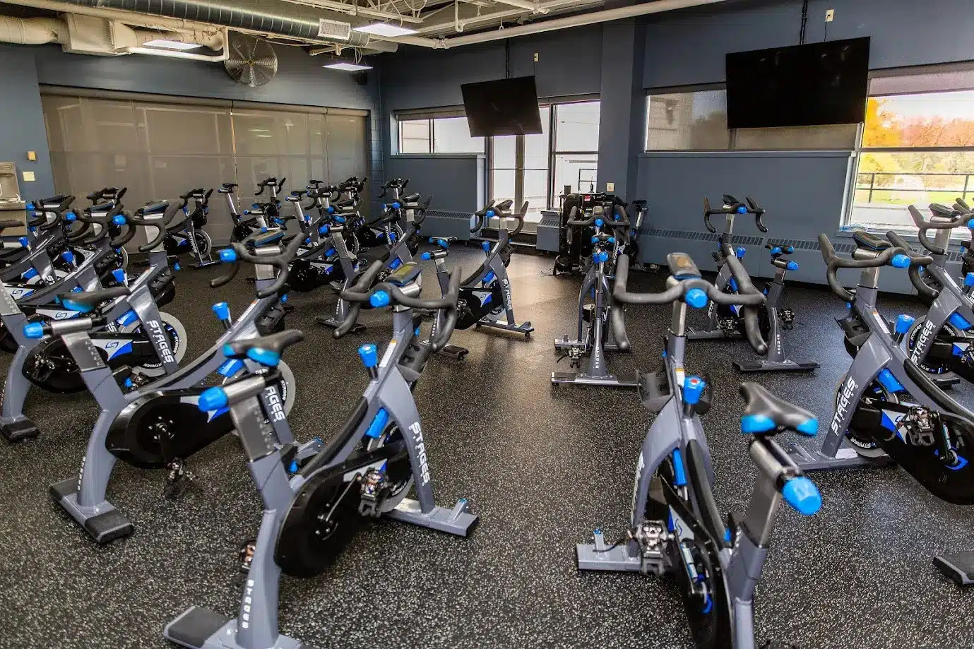 A room filled with multiple stationary bikes arranged in rows, each bike featuring a sleek design with blue accents. The room has large windows that let in natural light, and two large screens mounted on the walls. The flooring is a black speckled rubber material, and the room has a modern, clean, and well-lit atmosphere, suitable for group cycling classes.
