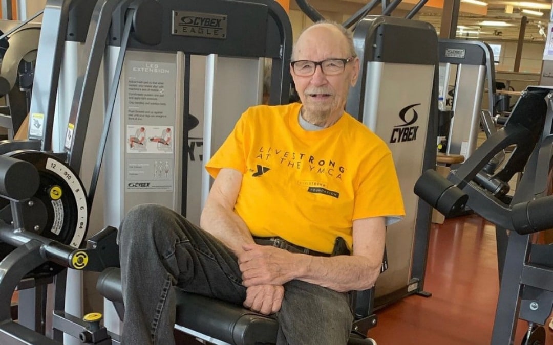 An older man sitting on gym equipment, wearing a yellow 'LIVESTRONG at the YMCA' shirt. He is smiling and appears to be in a fitness center, surrounded by exercise machines.