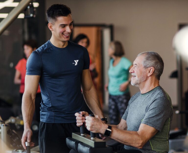 A young male trainer in a dark blue YMCA t-shirt is smiling and interacting with an older man who is using workout equipment at the YMCA gym. The older man is wearing a gray shirt and is also smiling. Other people can be seen exercising in the background, indicating a lively gym environment.