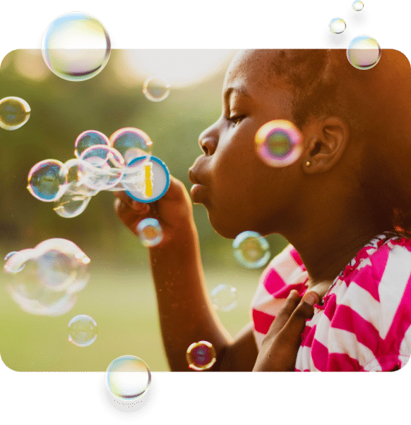A young girl in a pink and white striped dress is gently blowing soap bubbles in a sunny outdoor setting. The image captures the bubbles floating around her, reflecting shimmering colors. The girl is focused on the bubbles, with a serene expression on her face.