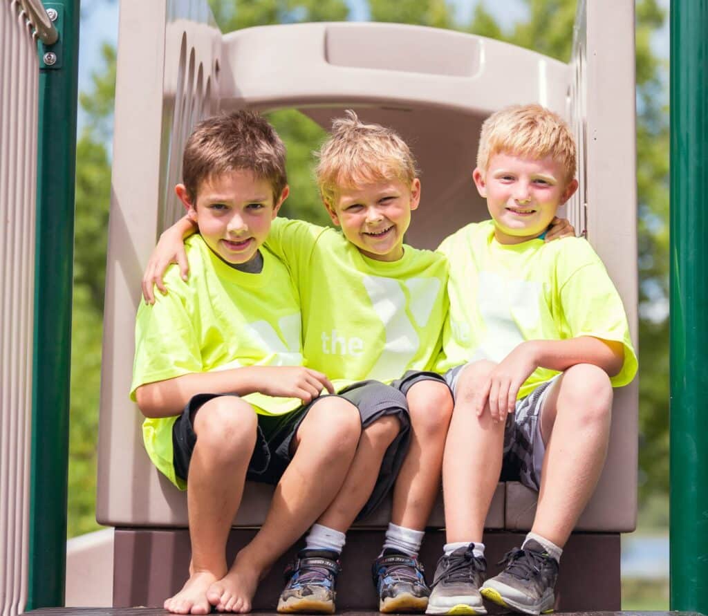 Three young boys, all wearing bright yellow YMCA t-shirts, sit together on a playground slide with their arms around each other, smiling happily at the camera. They are enjoying a sunny day outdoors, highlighting the camaraderie and fun of the YMCA's activities for children.