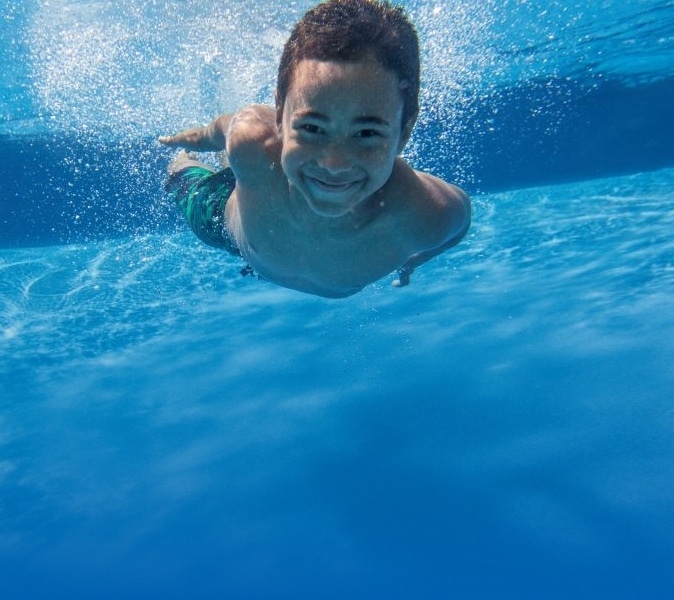 A young boy swimming underwater in a pool, smiling at the camera. He is wearing green swim trunks, and the water around him is clear and blue.