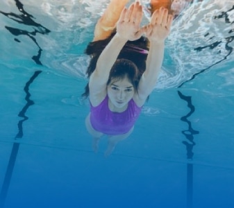 A woman swimming underwater in a pool, wearing a pink swimsuit. She has her arms extended in front of her, and the water around her is clear and blue