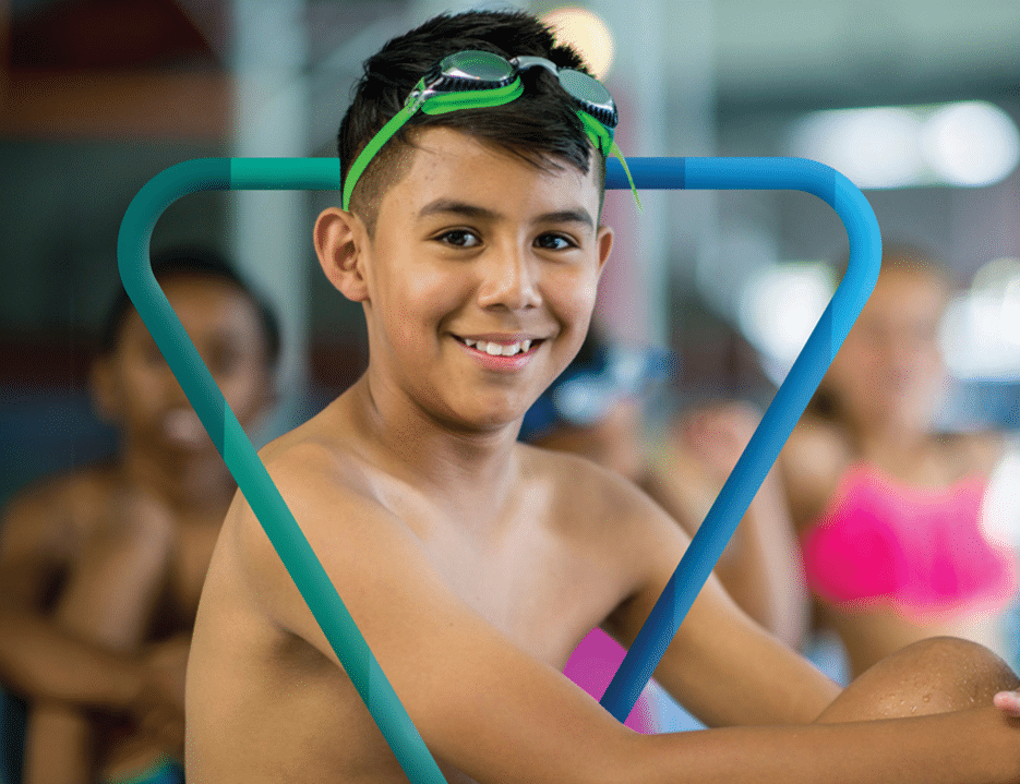 A young boy with a joyful smile poses at the poolside, wearing green swimming goggles on his head. He is framed by a stylized graphic of a blue 'Y' logo that curves around him, enhancing the vibrant, energetic atmosphere of the swimming area. Other children in swimwear are slightly blurred in the background, emphasizing the focus on the boy in the foreground.