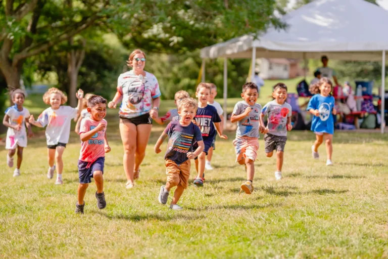 A joyful group of young children, guided by an adult, is running across a sunlit grassy field during a playful outdoor event. The children, displaying a variety of expressions of excitement and delight, are dressed in colorful summer clothes. The setting includes a large tent in the background, indicating a festive or community gathering.