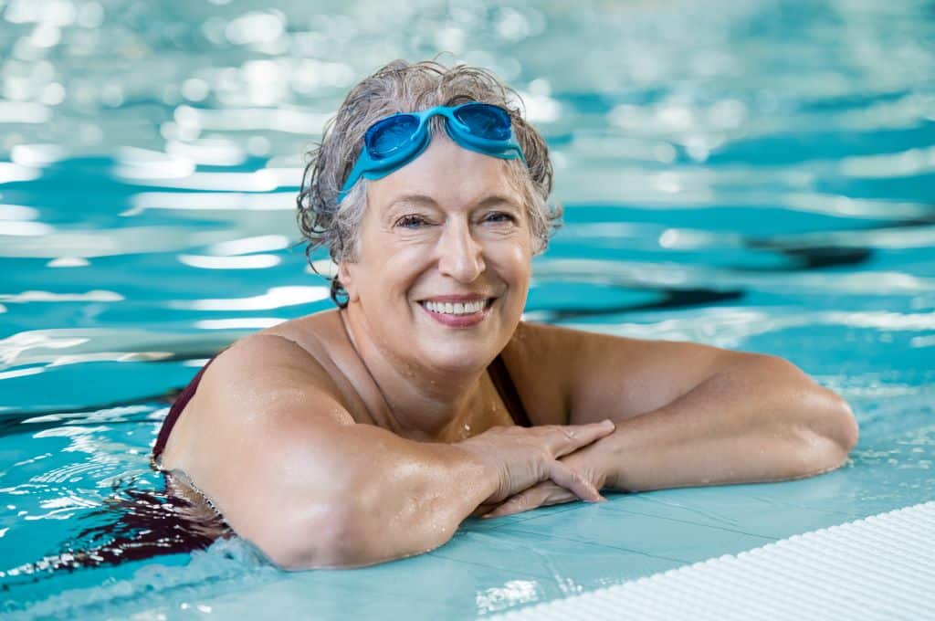 An elderly woman with short gray hair and blue swimming goggles resting on her forehead is smiling warmly while leaning on the edge of a swimming pool. The water around her is clear and inviting, reflecting the pool's gentle light. She appears relaxed and happy, enjoying her time in the pool.