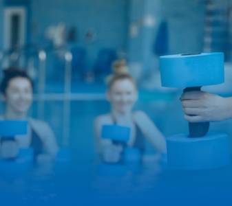 A group of female swimmers participating in an aqua aerobics class in a pool, holding blue foam dumbbells. The background is slightly blurred, focusing on the dumbbells in the foreground