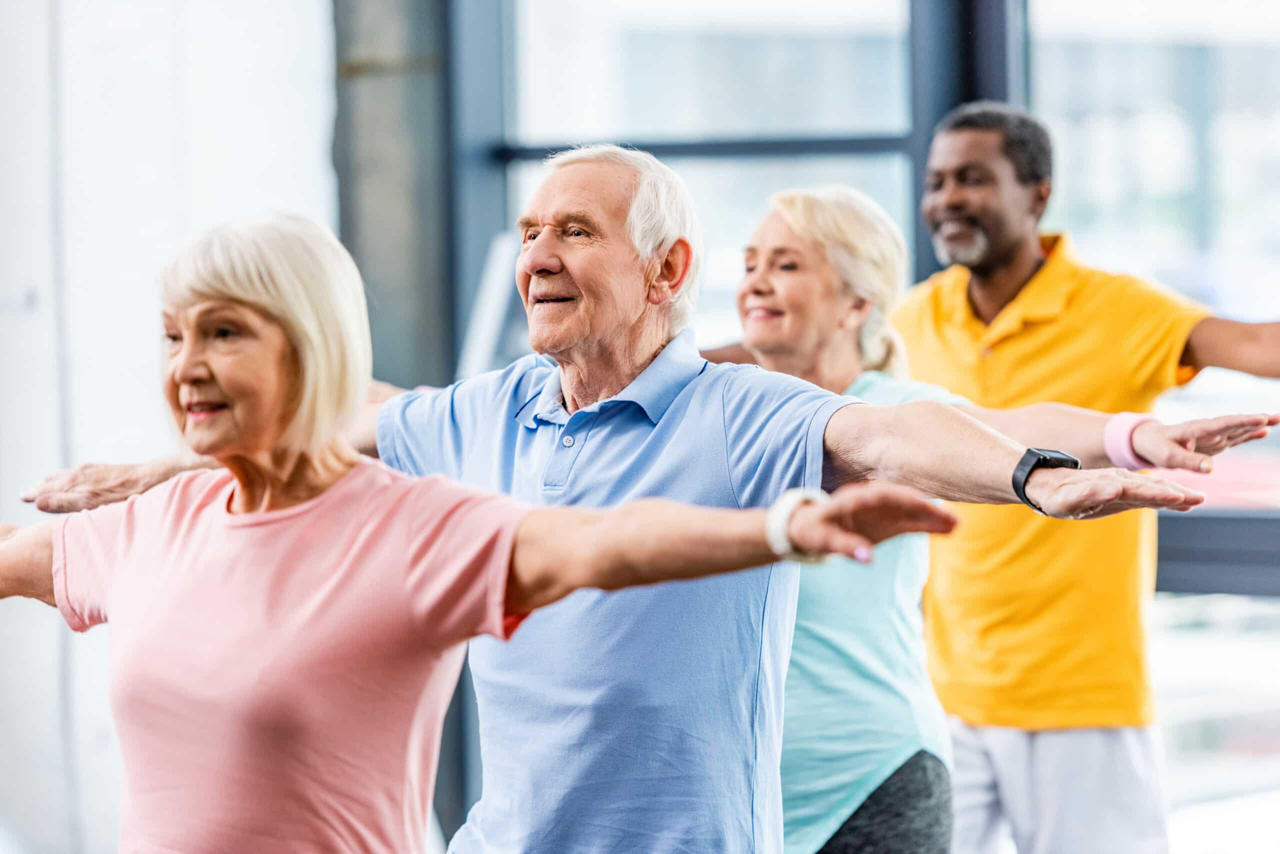 A group of happy seniors stretching their arms in a fitness class. They are standing in a line with arms extended, smiling, and wearing colorful workout clothes.