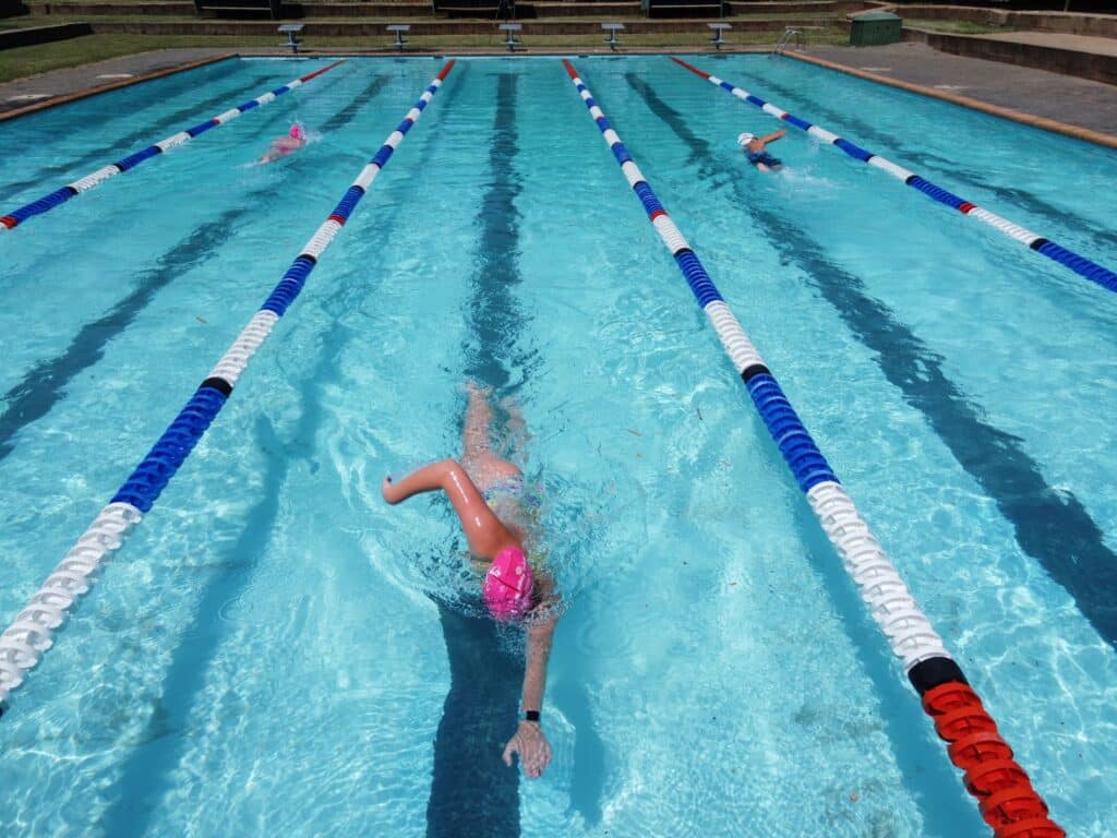 Overhead view of swimmers practicing in an outdoor pool, distinguished by crystal-clear turquoise waters divided by colorful lane markers. In the forefront, a swimmer wearing a pink swim cap is captured mid-stroke, demonstrating a powerful freestyle technique. Other swimmers are visible in adjacent lanes, each focused on their swim, in a scene depicting active training and competition preparation.