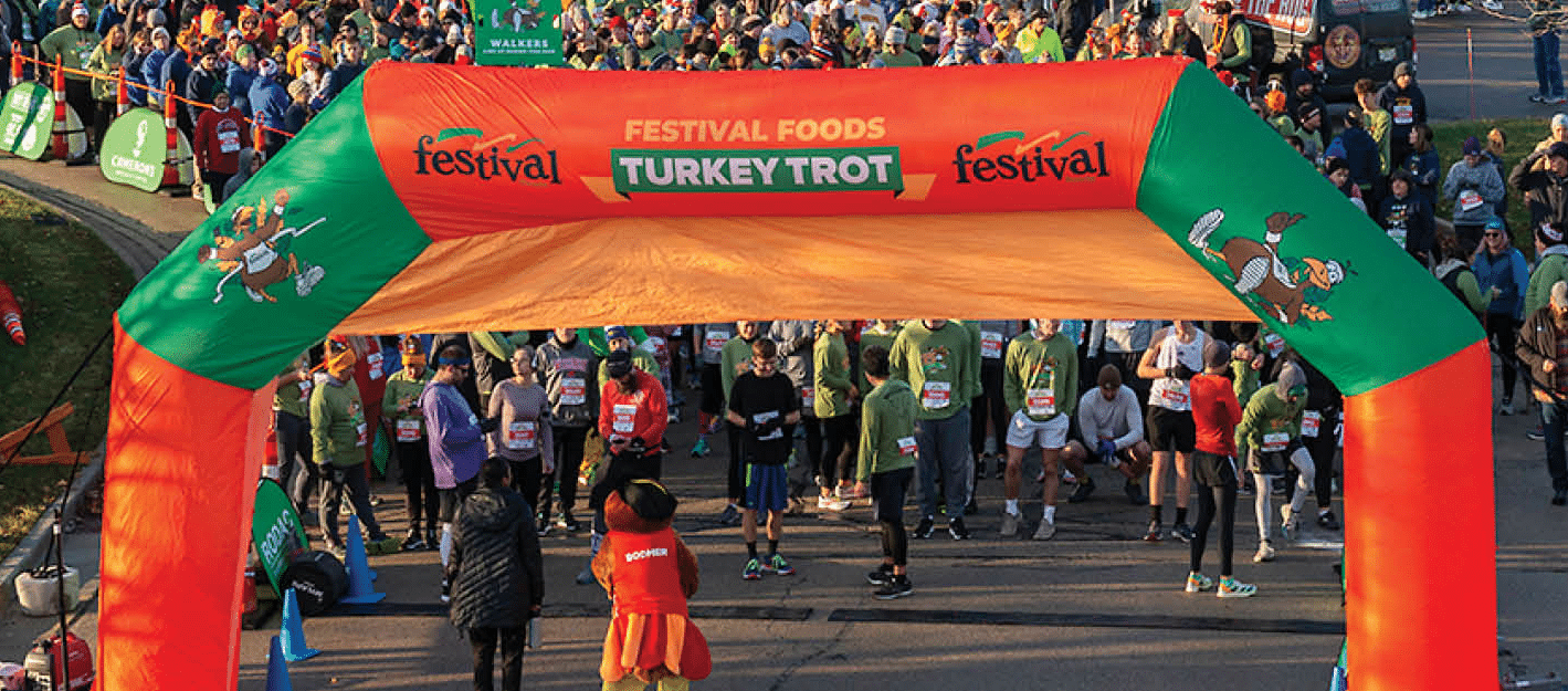 Participants at the start line of the Festival Foods Turkey Trot, gathering under a large orange and green inflatable arch decorated with turkey cartoons. The crowd, clad in various cold-weather athletic gear, appears eager and ready to begin the race. The scene is set in an urban area with early morning sunlight casting long shadows.