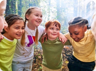 A group of four children are standing close together in a forest, smiling and with their arms around each other's shoulders. The girl on the left has brown hair and is wearing a light green shirt. Next to her is a girl with light hair in a white shirt, then a boy with red hair in a green shirt, and on the right is a girl with braided hair in a yellow shirt. The forest background is bright and lush, with sunlight filtering through the trees, creating a joyful and adventurous atmosphere.