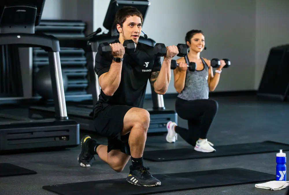 A male and a female gym trainer, both wearing black YMCA trainer uniforms, are performing lunges with dumbbells in a modern gym setting. The male trainer is in the foreground, demonstrating the exercise with focus, while the female trainer in the background mimics the motion with a confident smile. The gym is equipped with various cardio machines in the dimly lit background, emphasizing a professional training environment.