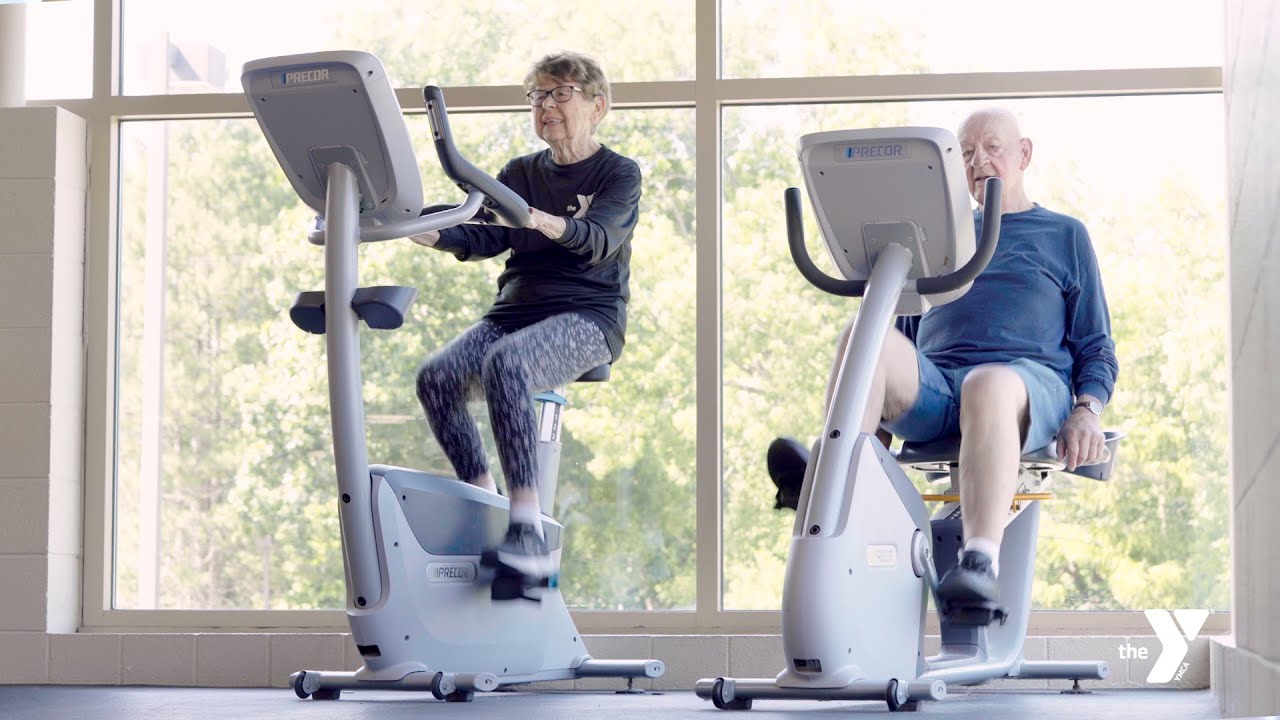 An elderly man and woman are exercising on stationary bikes in a gym with large windows, allowing natural light to fill the room. Both are dressed in comfortable workout clothes and appear focused on their exercise, demonstrating the YMCA's commitment to promoting fitness for all ages.