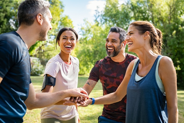 A diverse group of four adults is standing in a sunny park, smiling and laughing together. They appear to be friends or teammates, engaging in a cheerful conversation. The group consists of two men and two women, all dressed in casual athletic wear suitable for outdoor activities. One man is wearing a dark camouflage t-shirt, while another man wears a black t-shirt. One woman is wearing a light pink t-shirt with a black armband, and the other woman is dressed in a light blue tank top. They are standing close together, exuding camaraderie and joy.